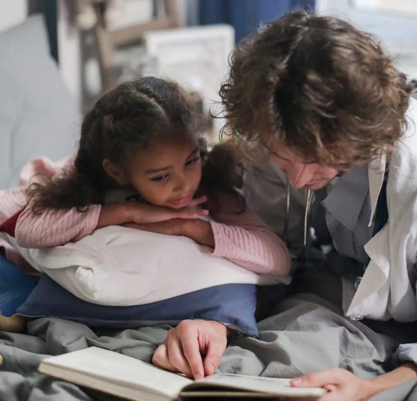A clinician reads a book with a young girl at her bedside at home